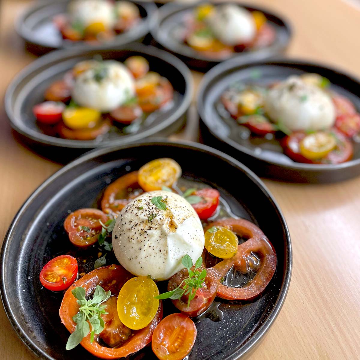 Five plates each feature a salad of sliced red and yellow heritage tomatoes, topped with a ball of burrata cheese, fresh herbs, black pepper, and a drizzle of olive oil, arranged on dark round plates.