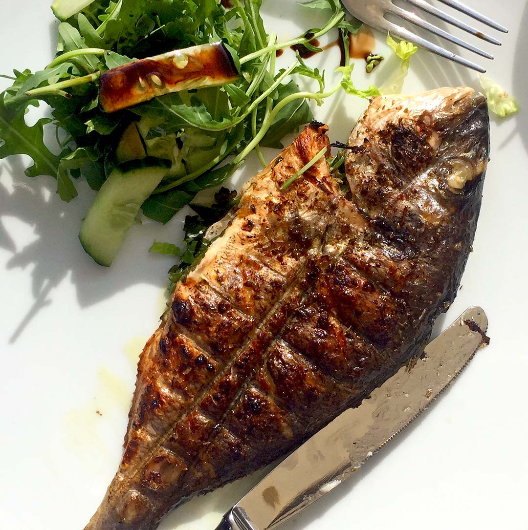 Grilled whole fish served on a white plate with a side of fresh green salad, including rocket and cucumber, accompanied by a fork and a knife.