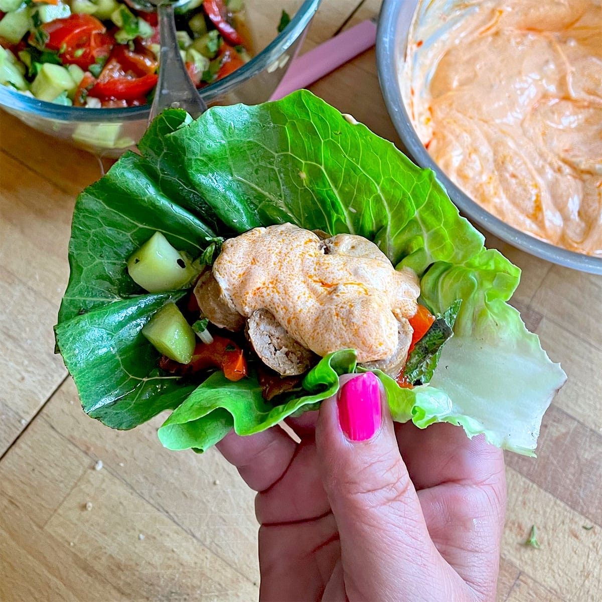 A hand with pink nail varnish holds a lettuce wrap filled with grilled chicken, diced vegetables, and topped with a dollop of orange creamy sauce. Bowls of salad and sauce are in the background on a wooden surface.