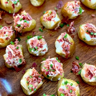 Bite-sized baked potato pieces topped with soured cream, crumbled bacon, and chopped chives are arranged on a wooden surface.
