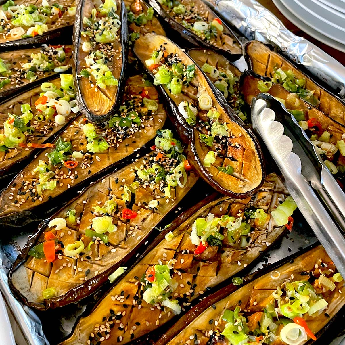 Roasted aubergine halves topped with chopped spring onions, red chilli, sesame seeds, and herbs are arranged on a tray lined with foil. Metal tongs are placed on the right side of the tray.