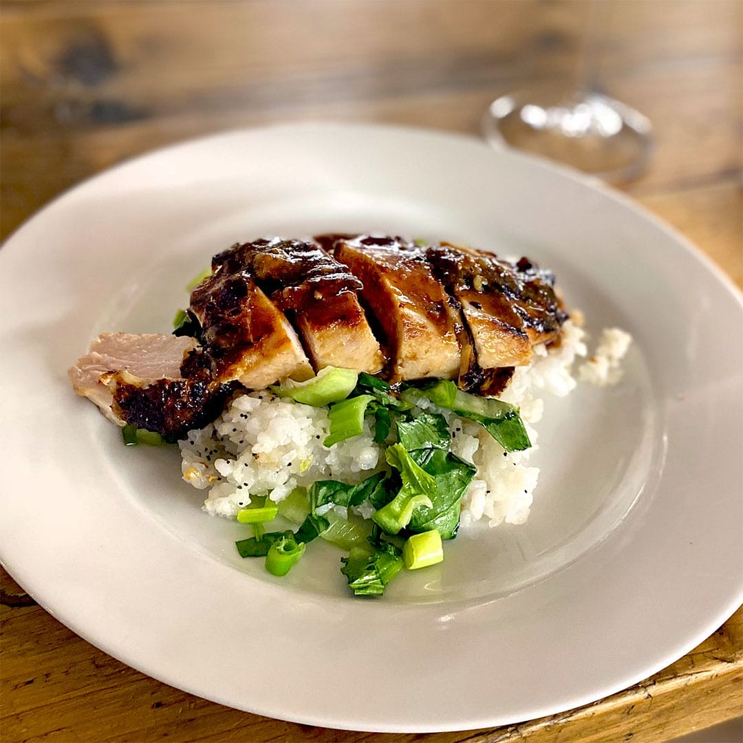 Sliced grilled chicken breast served on a bed of white rice and chopped green vegetables, arranged on a white plate on a wooden table. A blurred wine glass is visible in the background.