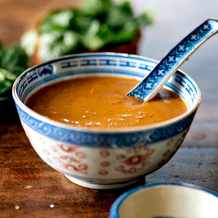 A blue-and-white patterned bowl filled with brown peanut sauce, with a matching spoon resting inside. The bowl sits on a wooden surface, and green herbs are blurred in the background.
