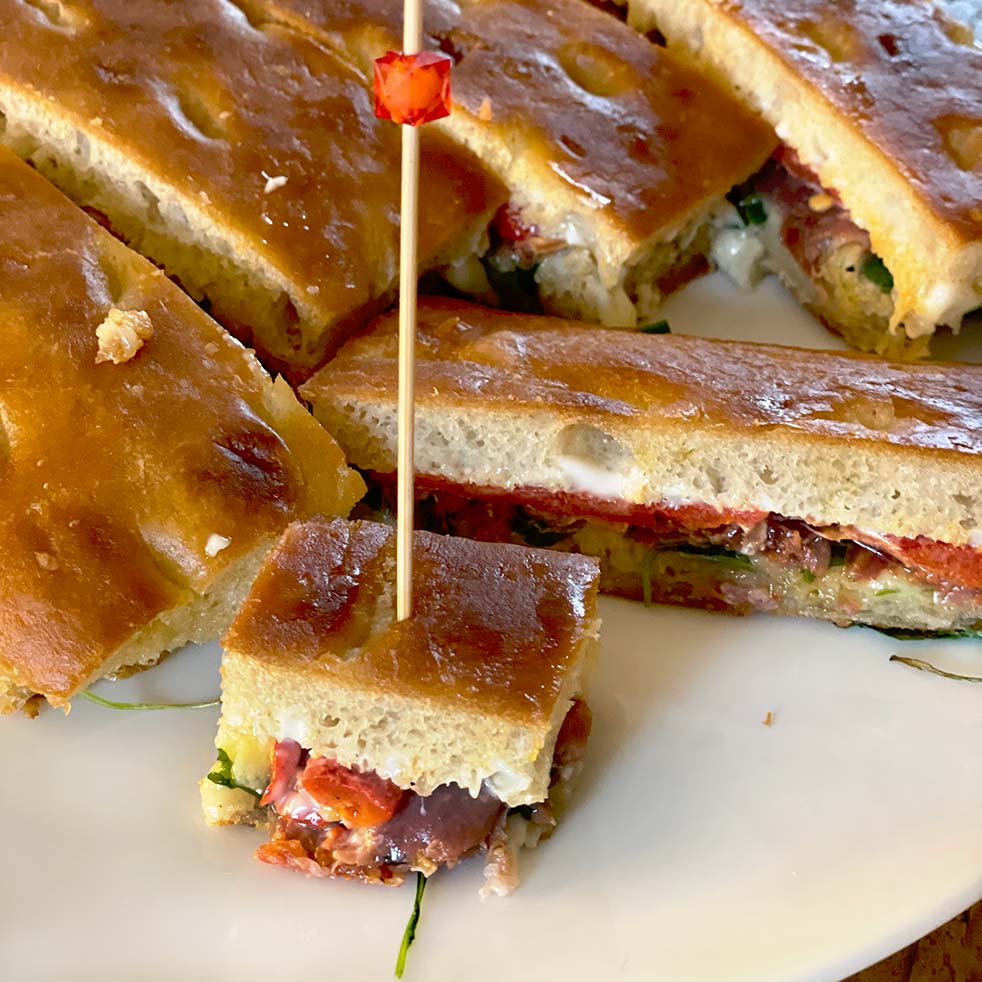 A close-up of sliced focaccia sandwiches with fillings such as vegetables and cheese, served on a white plate. One small sandwich piece is secured with a cocktail stick.