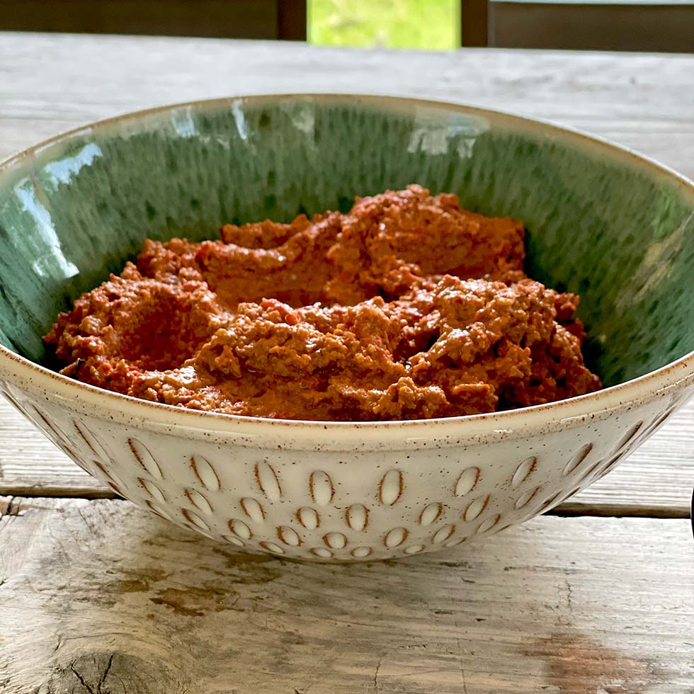 A decorative ceramic bowl filled with a chunky, reddish-brown dip sits on a wooden table, with a blurred outdoor scene in the background.
