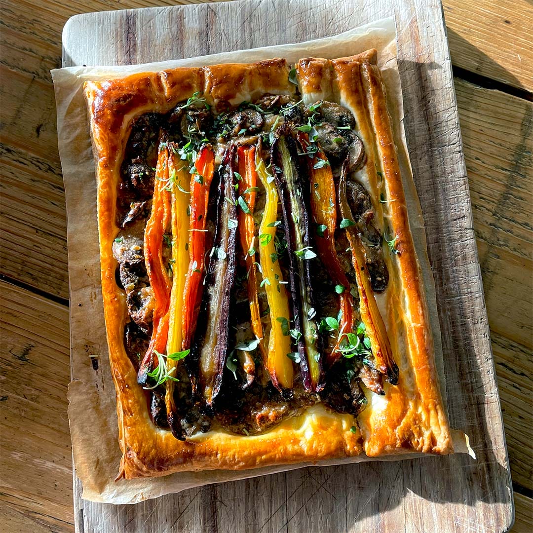 A rectangular vegetable tart with flaky golden pastry, topped with colourful roasted carrots, herbs, and a dark spread, sits on baking parchment on a wooden chopping board in sunlight.