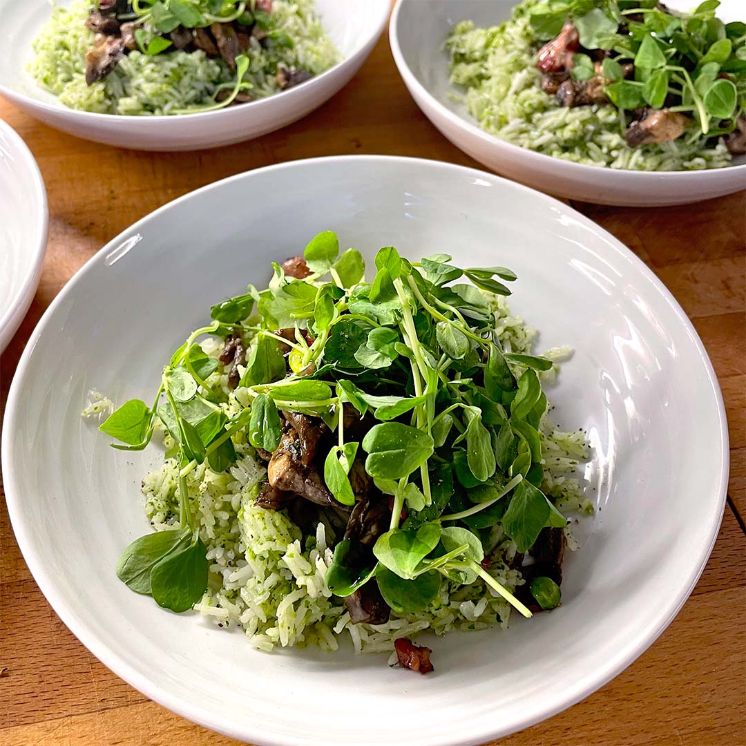 A white bowl filled with green herb rice, topped with sautéed mushrooms and fresh leafy microgreens, sits on a wooden table with similar bowls in the background.