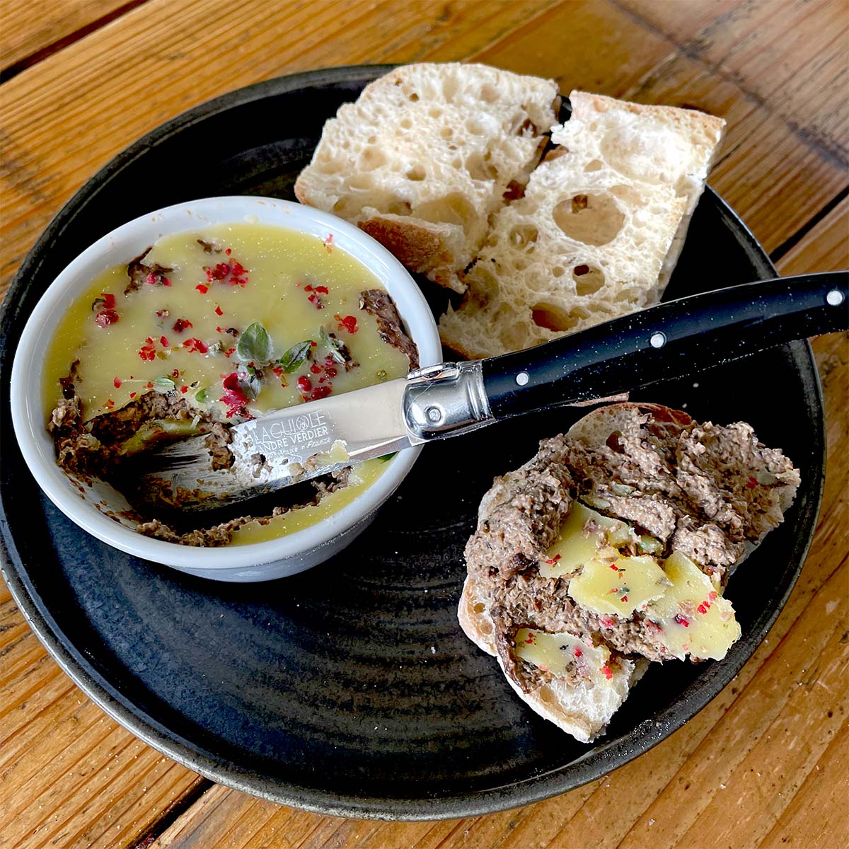 A black plate with a ramekin of pâté topped with herbs and spices, slices of rustic bread, and a knife spread with pâté and butter on a wooden table.
