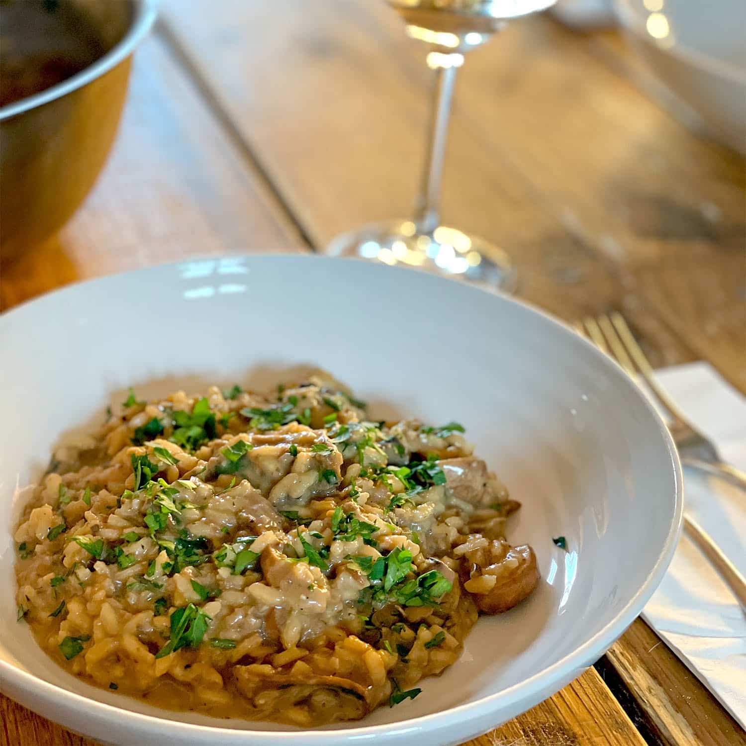 A white bowl of creamy mushroom risotto garnished with chopped parsley sits on a wooden table beside a glass of white wine, with a fork and serviette nearby.