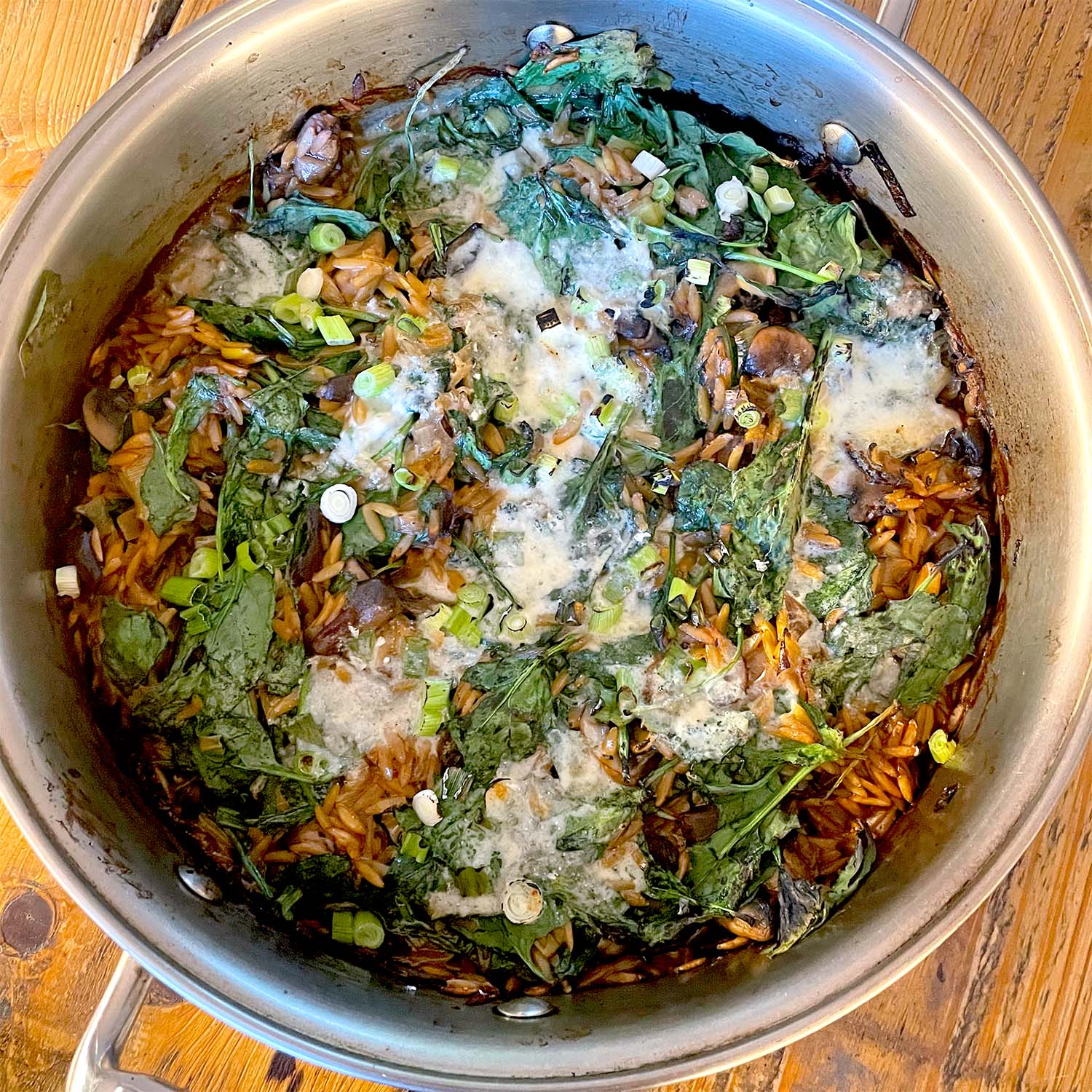 A pot of cooked orzo pasta mixed with spinach, mushrooms, spring onions, and melted cheese, sitting on a wooden table. The ingredients are combined and lightly browned on top.