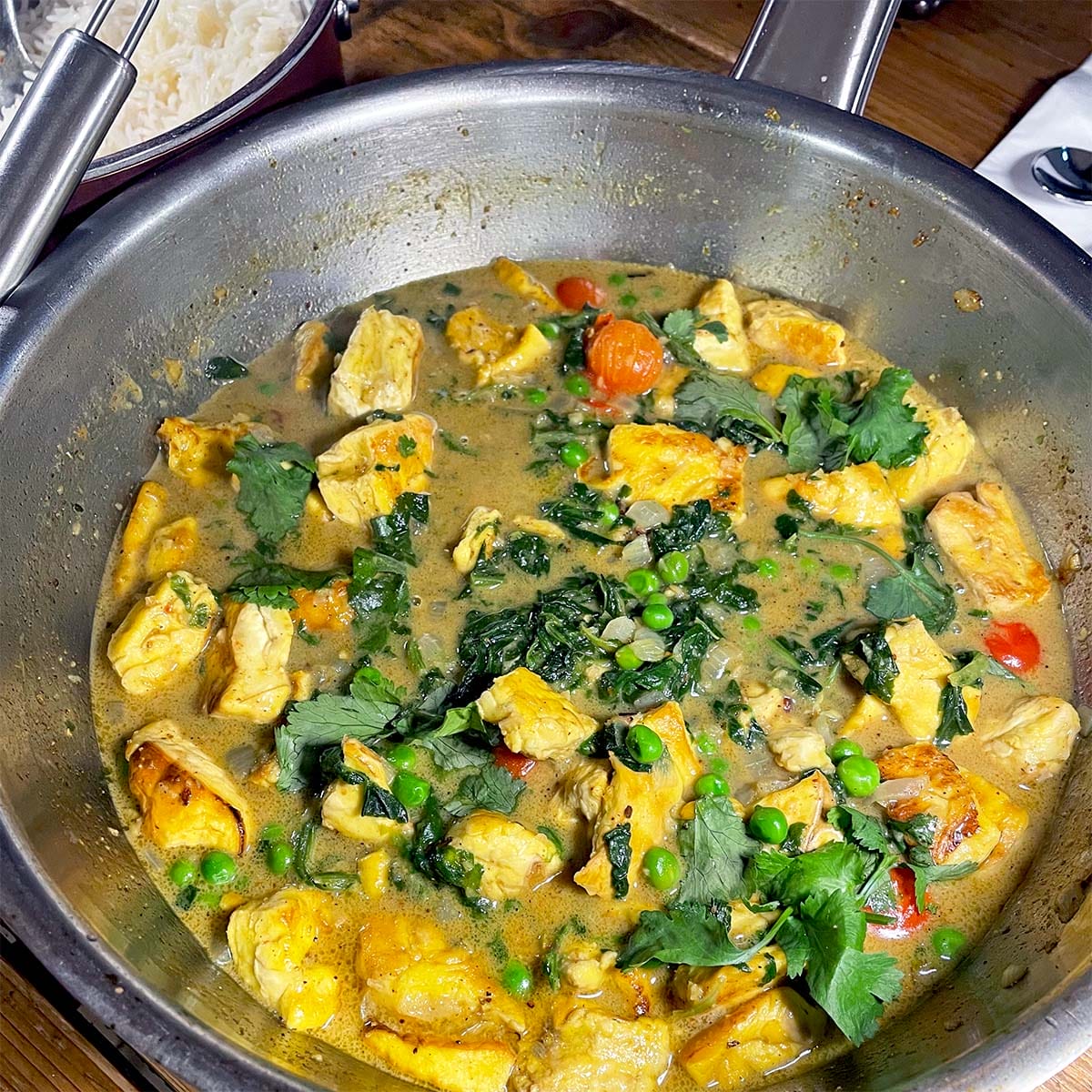 A stainless steel pan filled with a chicken curry dish, featuring chunks of chicken, peas, cherry tomatoes, fresh coriander, and a creamy sauce. A bowl of white rice is visible in the background.