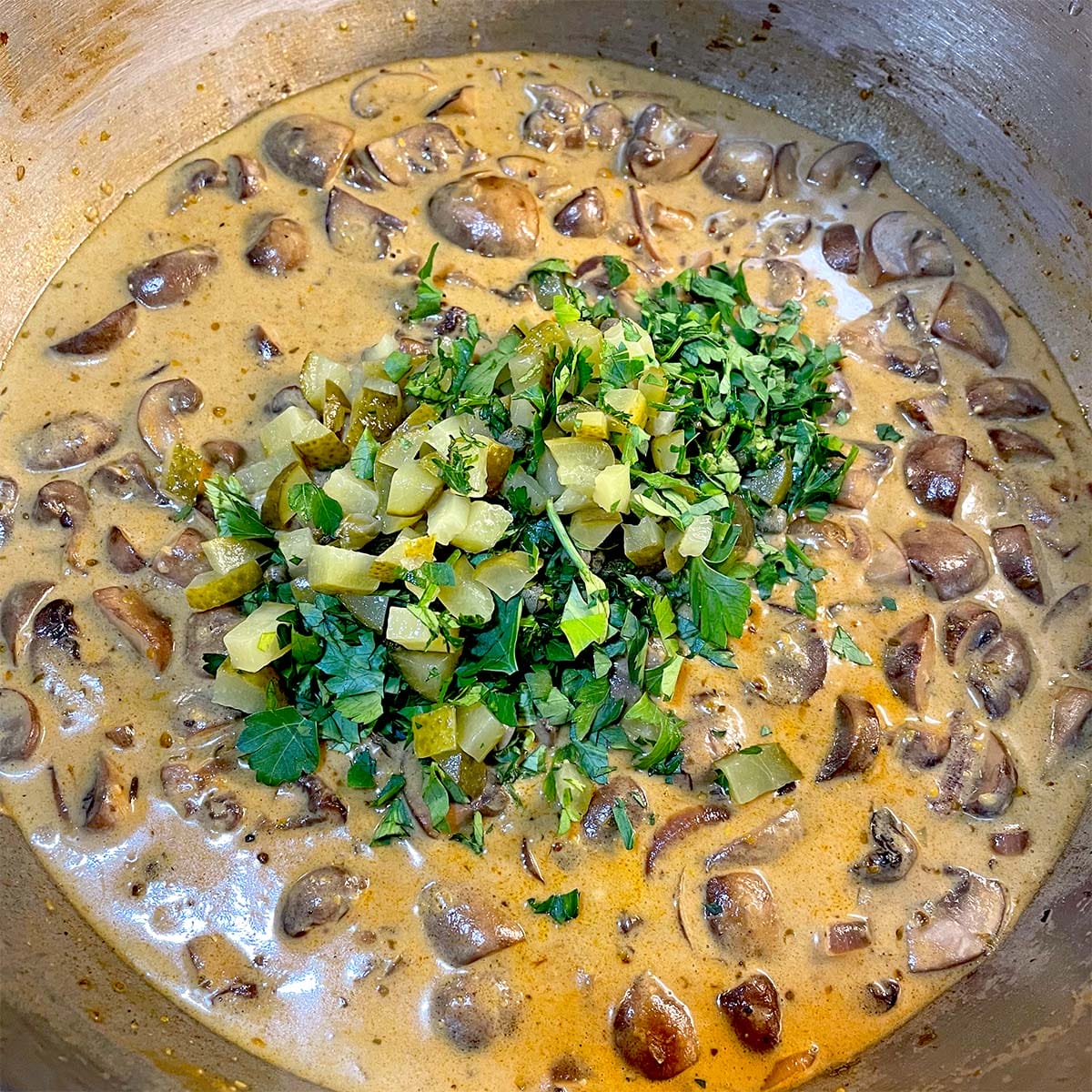 A creamy mushroom stew in a saucepan, topped with chopped gherkins and fresh parsley.