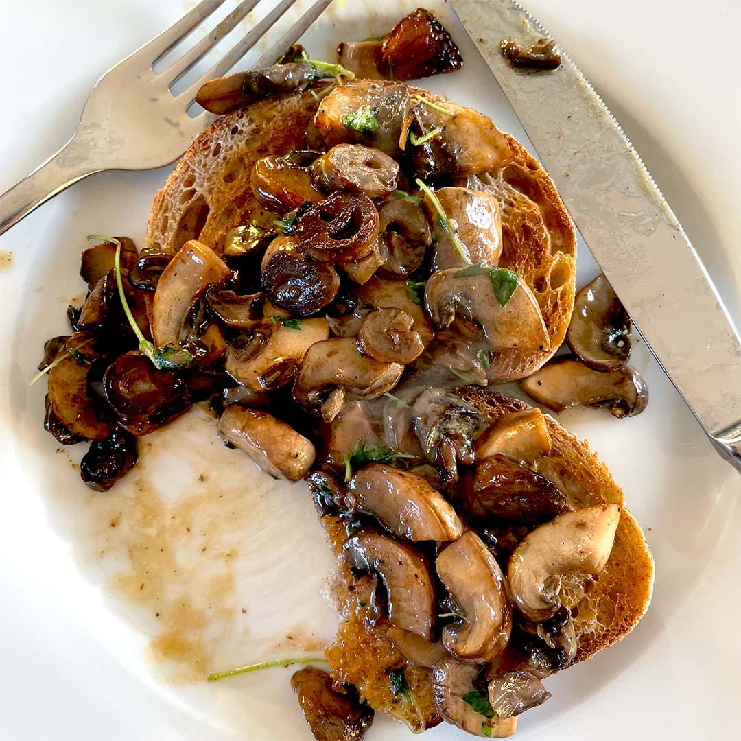 Two slices of toasted bread topped with sautéed mushrooms and herbs on a white plate, with a fork and knife placed beside the food.