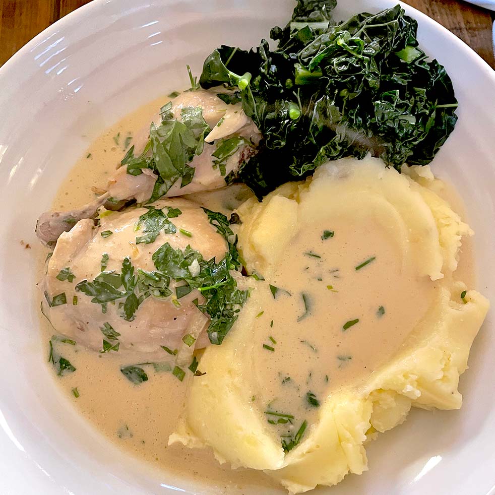 A plate with creamy mashed potatoes, chicken in herb sauce, and a serving of cooked dark leafy greens, all garnished with chopped fresh herbs.