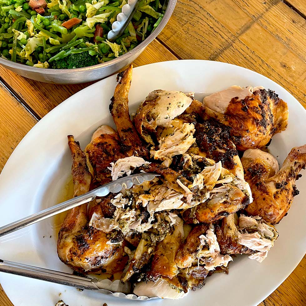 A platter of roasted, carved chicken with metal tongs, served on a wooden table next to a bowl of fresh green salad with broccoli and nuts.