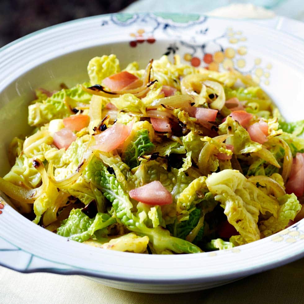 A decorative bowl filled with a fresh salad made of chopped leafy greens, caramelised onions, and diced pink vegetables, possibly apples or tomatoes. The salad is lightly tossed and appears crisp and colourful.