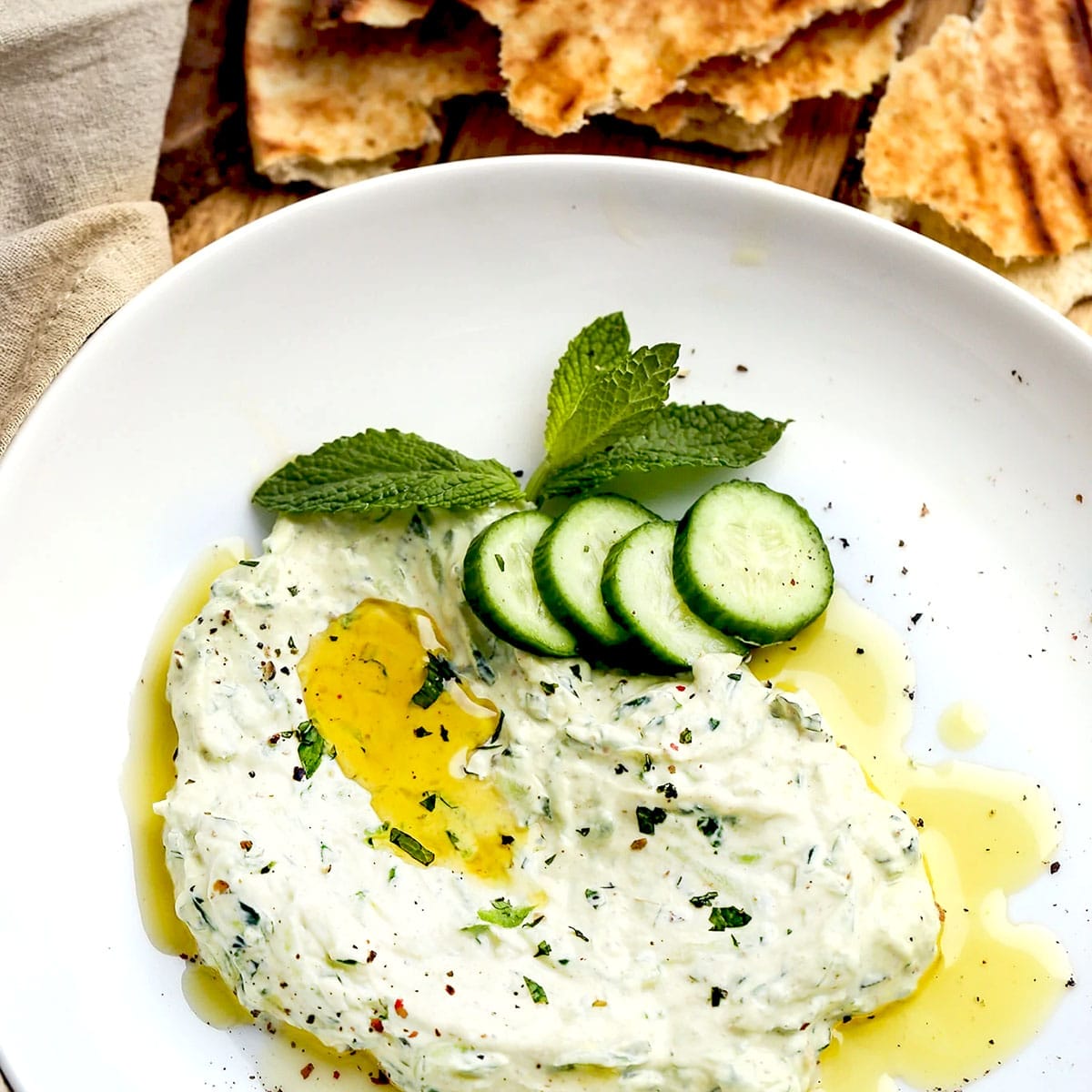 A white plate with creamy tzatziki dip, garnished with olive oil, mint leaves, and cucumber slices. Pieces of flatbread are in the background.