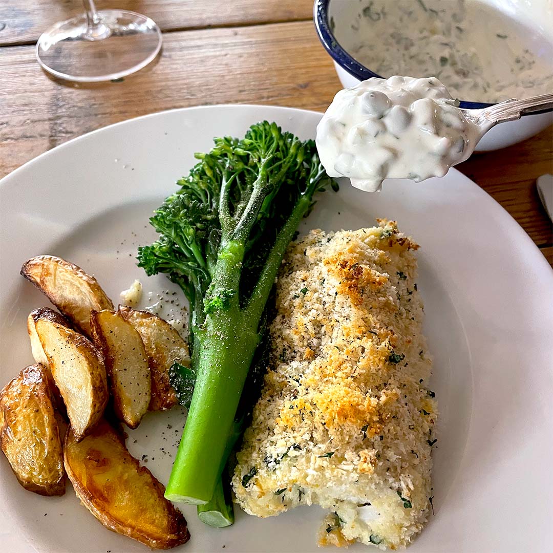 A plate with baked fish topped with breadcrumbs, roasted potato wedges, and steamed tenderstem broccoli. A spoonful of creamy tartare sauce is being served from a small bowl.