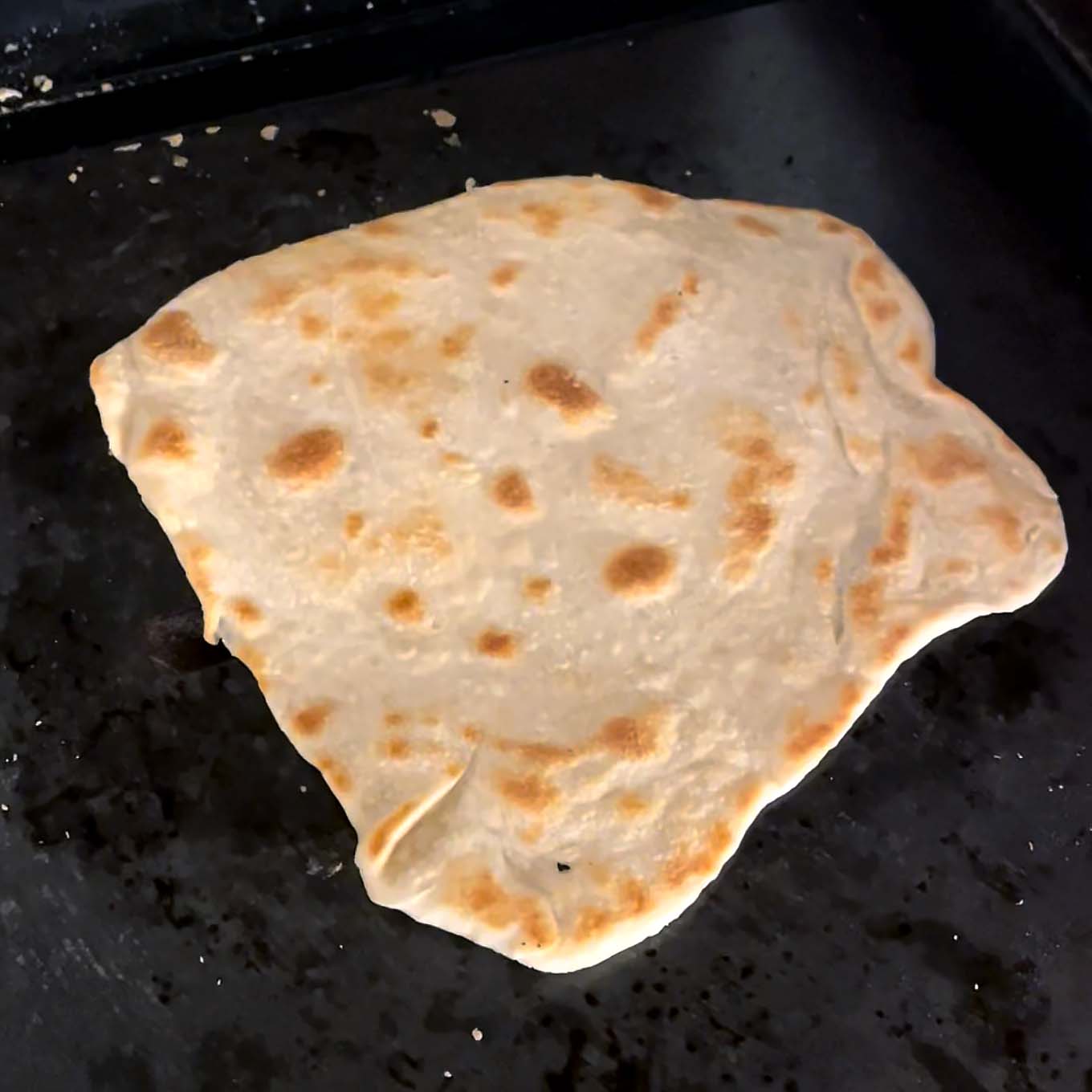 A piece of unevenly shaped flatbread with golden-brown spots cooks on a dark, oily griddle.
