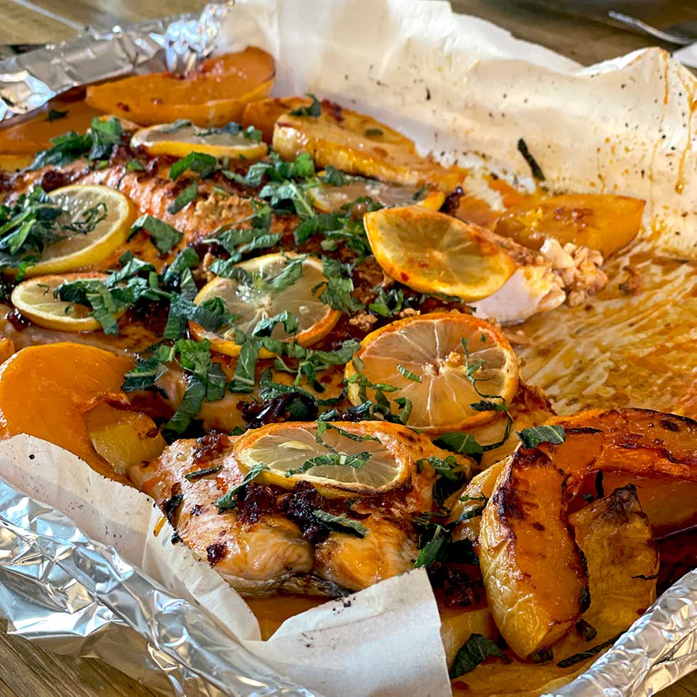 Baked salmon fillet topped with lemon slices and fresh herbs, surrounded by roasted orange and yellow vegetables on a foil-lined baking tray.