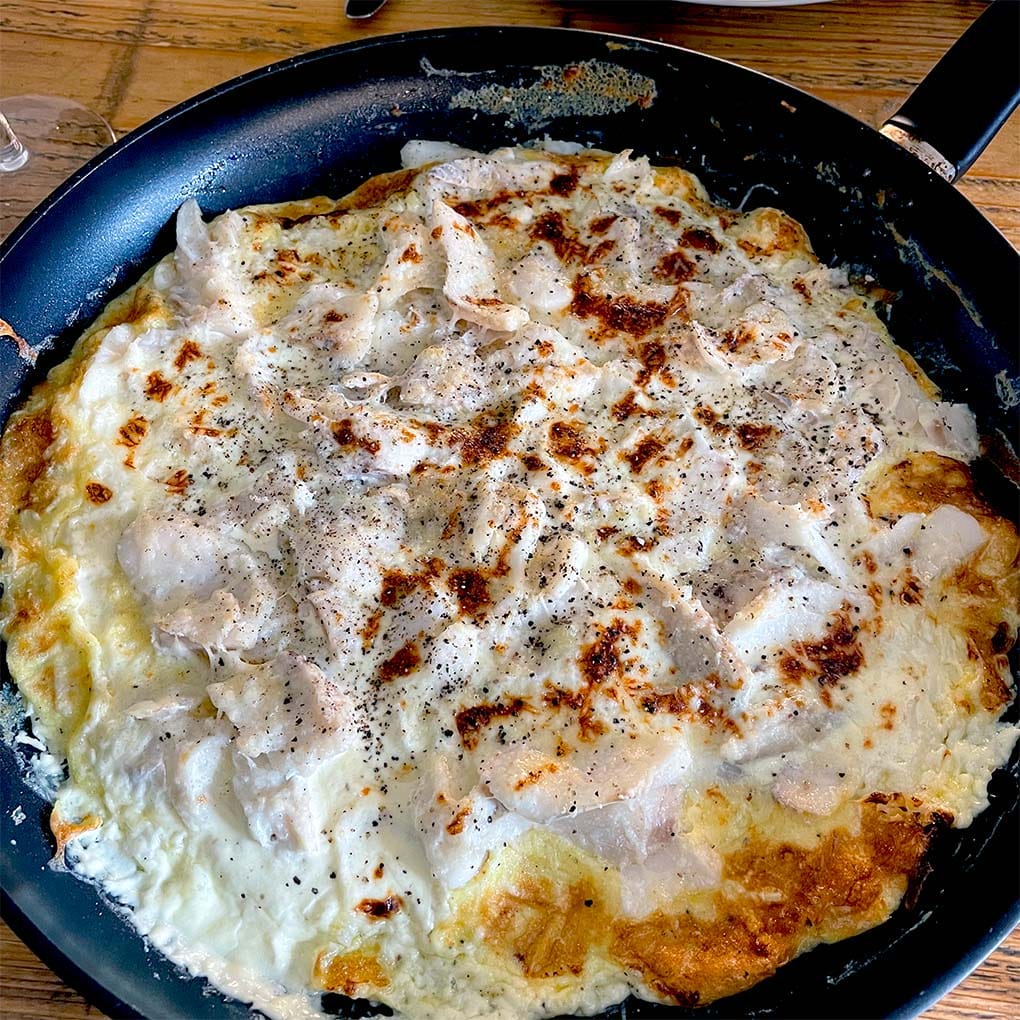 A black frying pan on a wooden table contains a cooked, golden-brown omelette topped with melted cheese and black pepper. The omelette appears fluffy with slightly crispy edges.