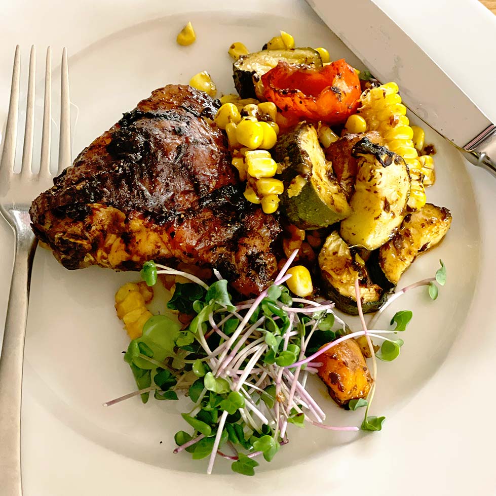 A plate with grilled chicken, roasted vegetables including courgette, tomato, and sweetcorn, garnished with fresh microgreens. A fork and knife are placed beside the food on the white plate.