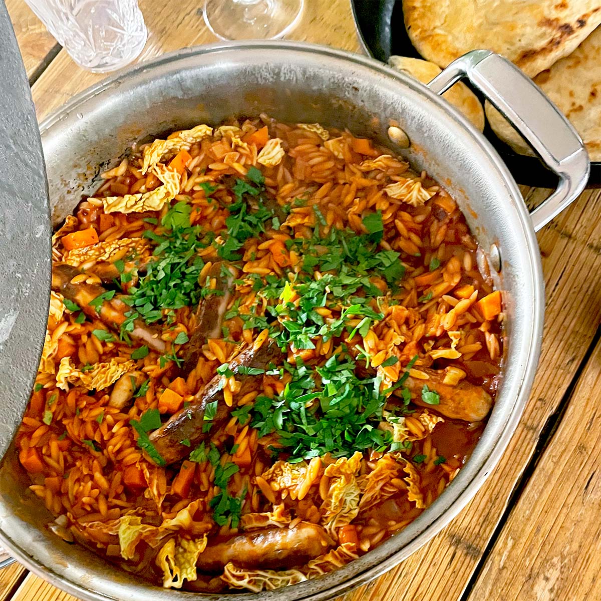 A large saucepan filled with a stew containing sausages, vegetables, orzo pasta, and chopped herbs on top. Flatbread and a glass are placed on the wooden table beside the saucepan.