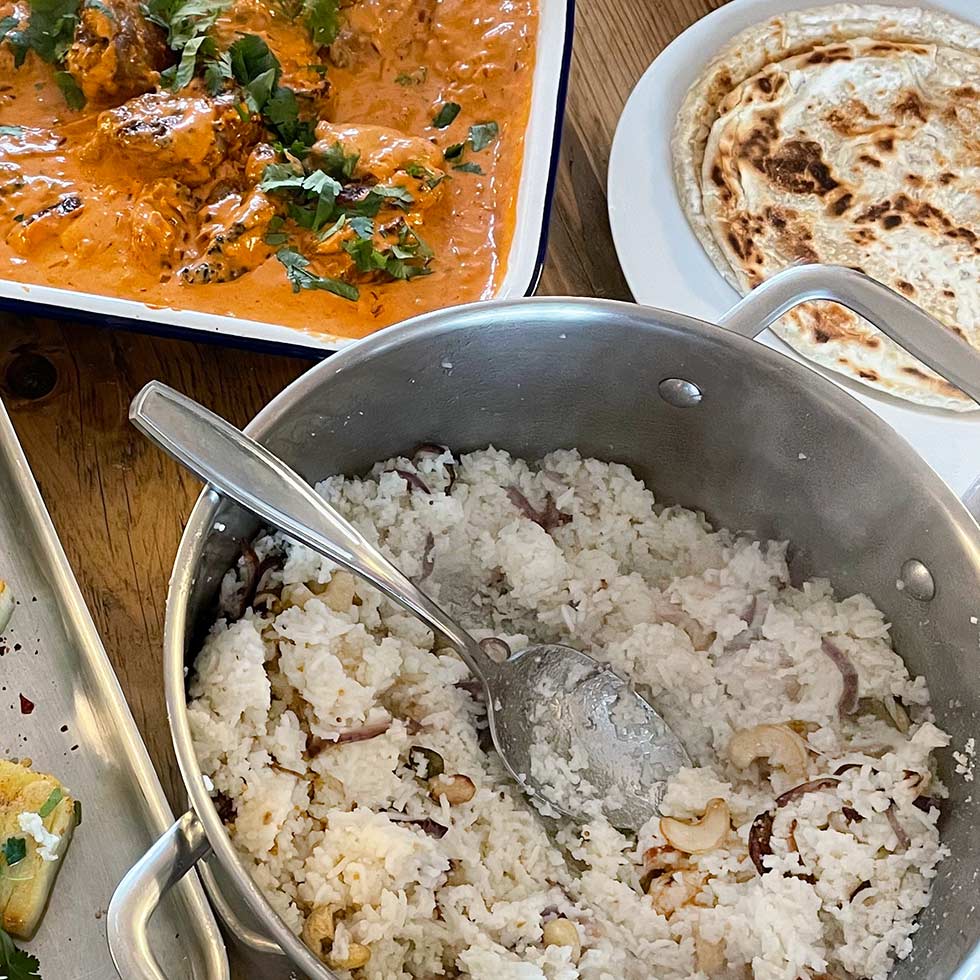 A pot of cooked white rice with cashews and onions sits next to a dish of creamy orange curry garnished with coriander and a plate of round flatbread on a wooden table.