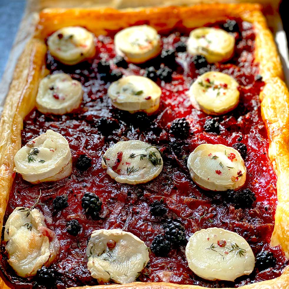 Rectangular puff pastry tart topped with a red jam-like layer, blackberries, and round slices of goats' cheese, garnished with herbs and pink peppercorns. The crust is golden and flaky.