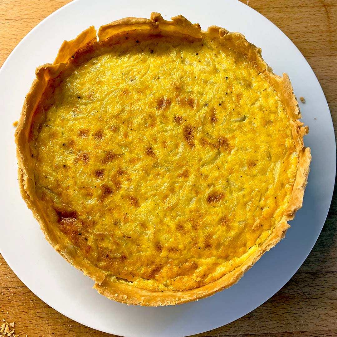 A golden-brown quiche with a flaky pastry sits on a white plate atop a wooden surface. The filling appears smooth and slightly browned on top.