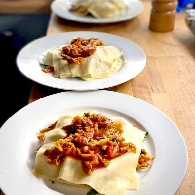 Three white plates sit on a wooden counter, each with homemade lasagne featuring visible pasta sheets and a chunky tomato-based sauce with meat and vegetables, ready to be served.