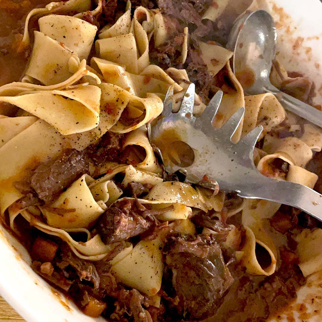 Wide ribbons of pasta tossed with chunks of braised beef in a rich, dark sauce, served in a white dish with a pasta server and metal spoon.