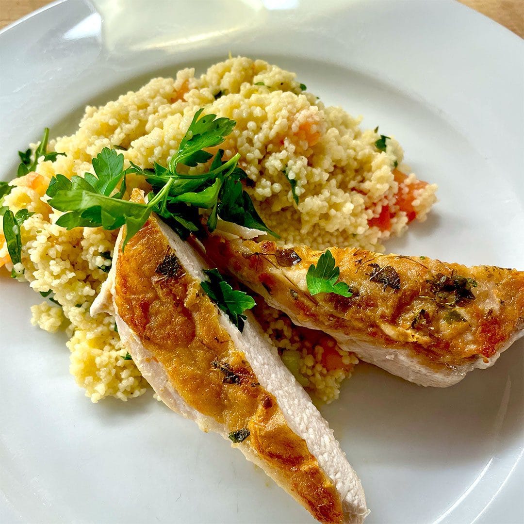 A white plate with grilled chicken breast slices garnished with parsley, served alongside a mound of couscous mixed with chopped vegetables.