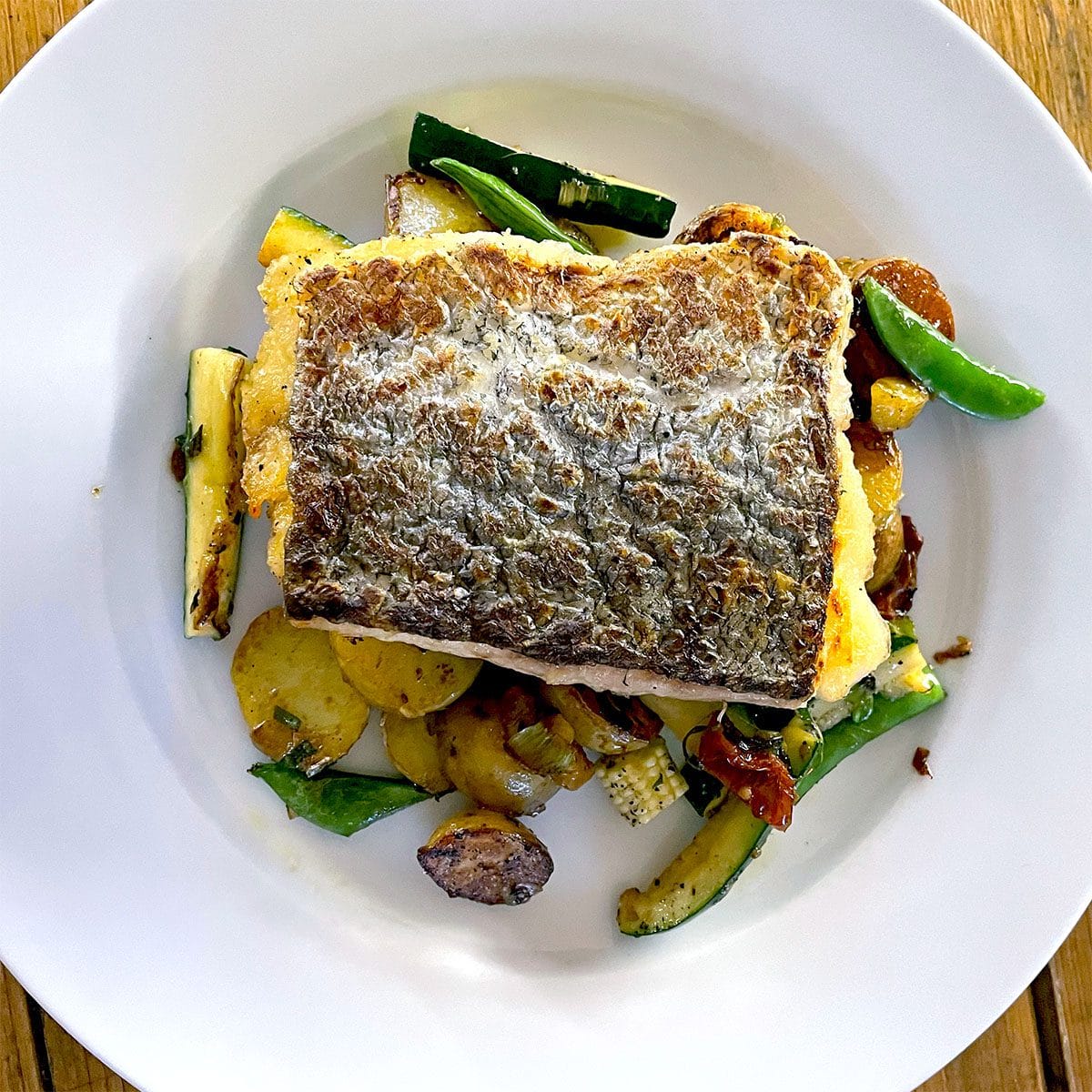 A fillet of fish with crispy skin served on sautéed vegetables, including courgette, potatoes, and green beans, on a white plate.
