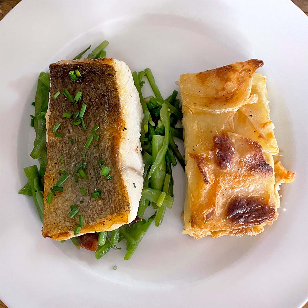 A white plate with a piece of fish with crispy skin and chopped chives, served on green beans, next to a slice of cheesy potato gratin.
