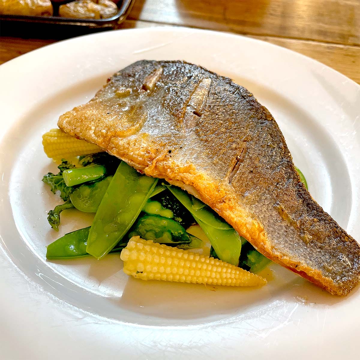 A cooked fish fillet with crispy skin served on a bed of baby corn, broccoli, and mangetout, presented on a white plate.