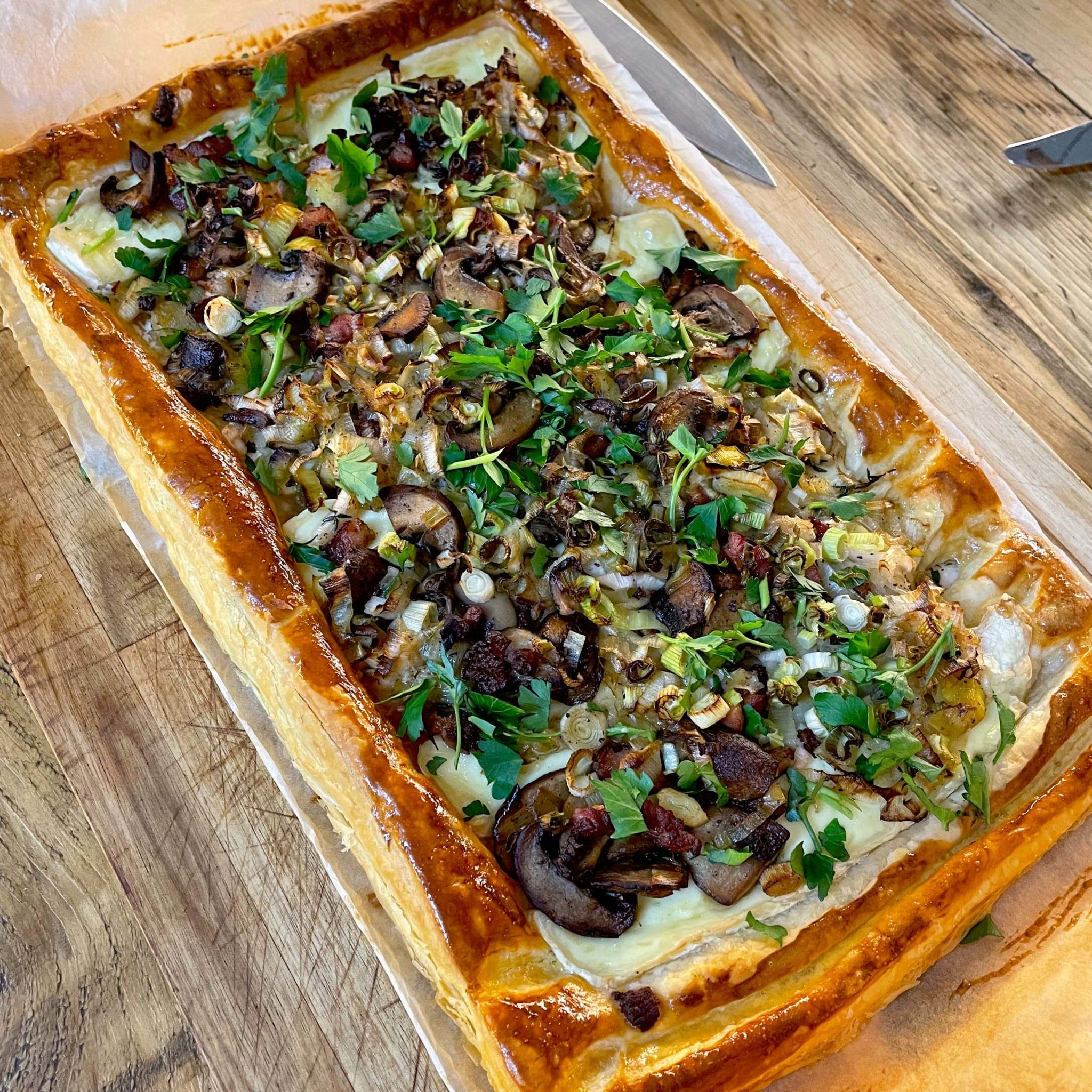 A rectangular puff pastry tart topped with sliced mushrooms, fresh herbs, and onions sits on baking parchment on a wooden surface. A knife is visible nearby.