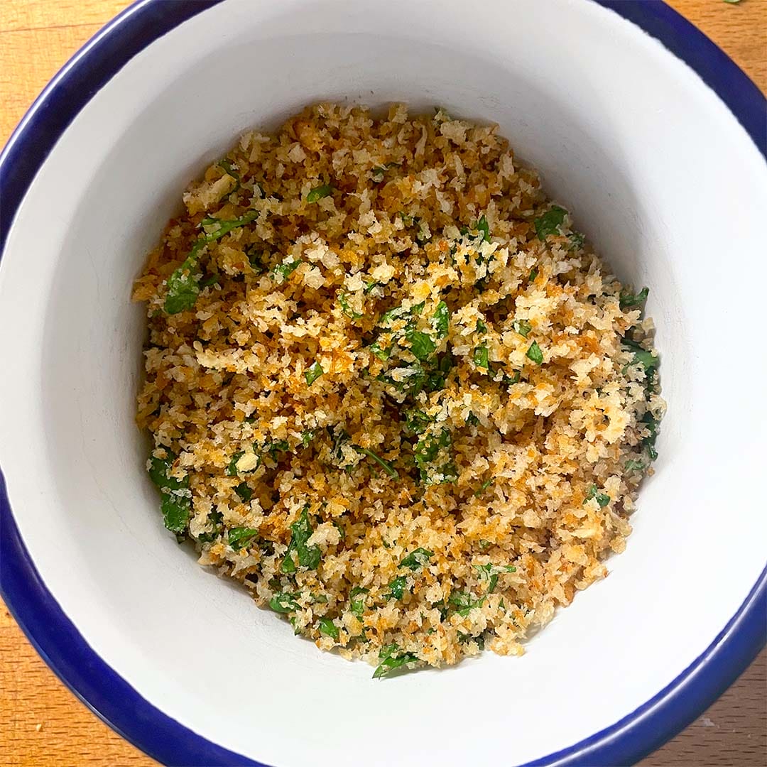 A white bowl with a blue rim filled with golden-brown seasoned breadcrumbs mixed with chopped green herbs, sitting on a wooden surface.