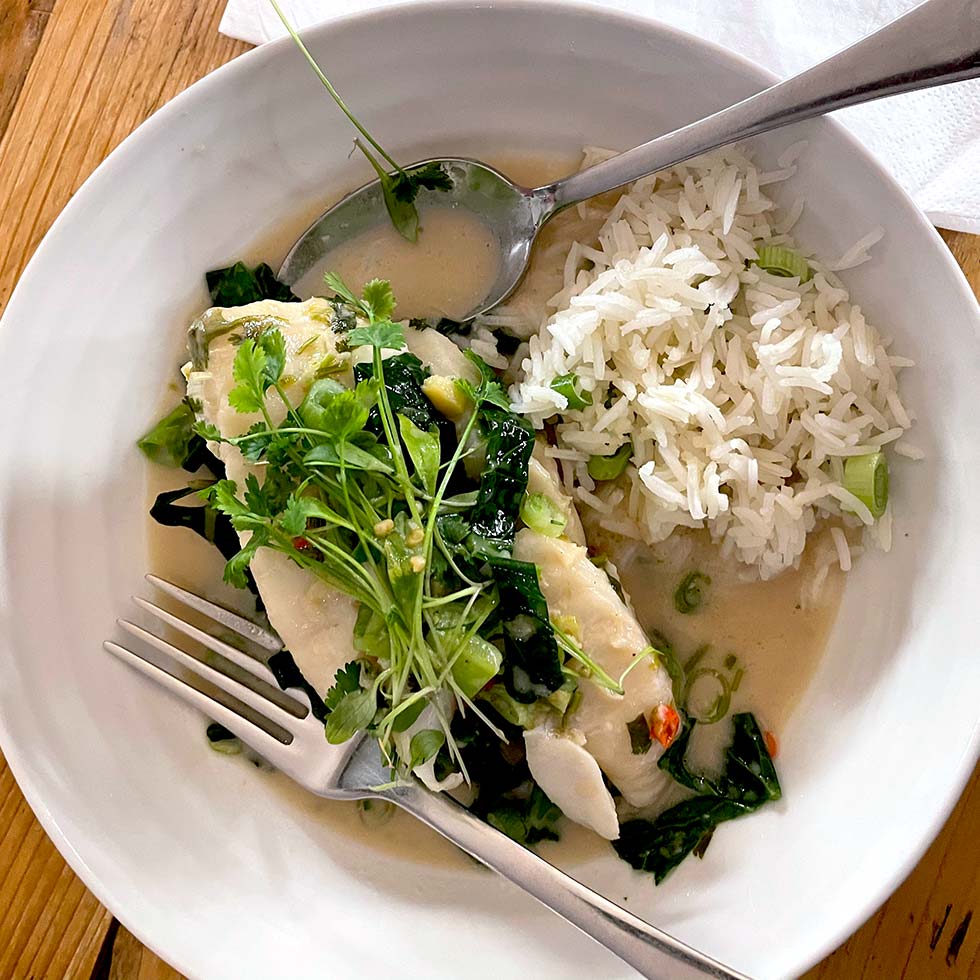 A white bowl with steamed white fish topped with fresh herbs and greens in a light sauce, served alongside a portion of white rice with spring onions. A fork and spoon rest on the edge of the bowl.
