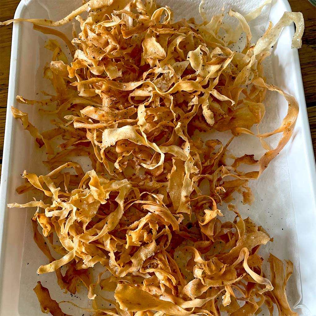 A tray lined with kitchen paper holds a pile of crispy, golden-brown parsnip crisps, freshly cooked and lightly seasoned.