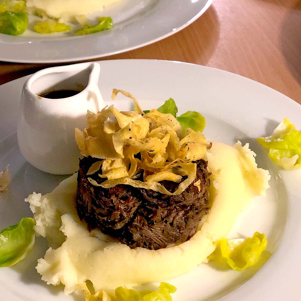 A plated dish featuring shredded beef topped with crispy fried onions, surrounded by creamy mashed potato and garnished with green leaves, with a small jug of brown sauce on the side.