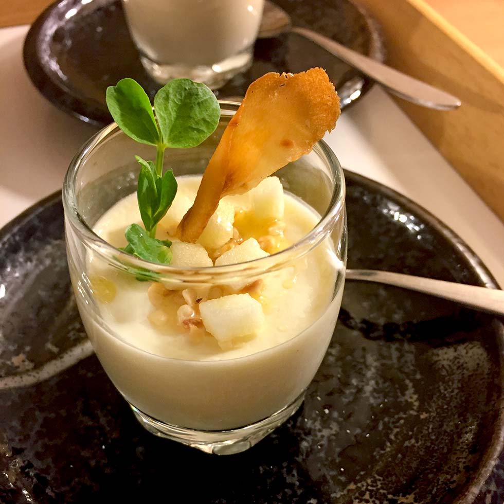 A glass dessert dish filled with creamy panna cotta, topped with diced fruit, a crunchy garnish, and a small green sprig, placed on a dark plate with a spoon beside it.