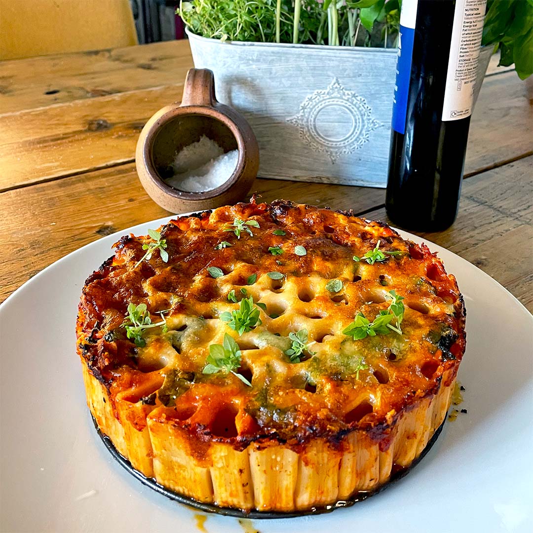 A baked pasta pie with a golden, cheesy top and herbs is served on a white plate. There’s a wooden table, a salt pot, a bottle, and a planter with greens in the background.
