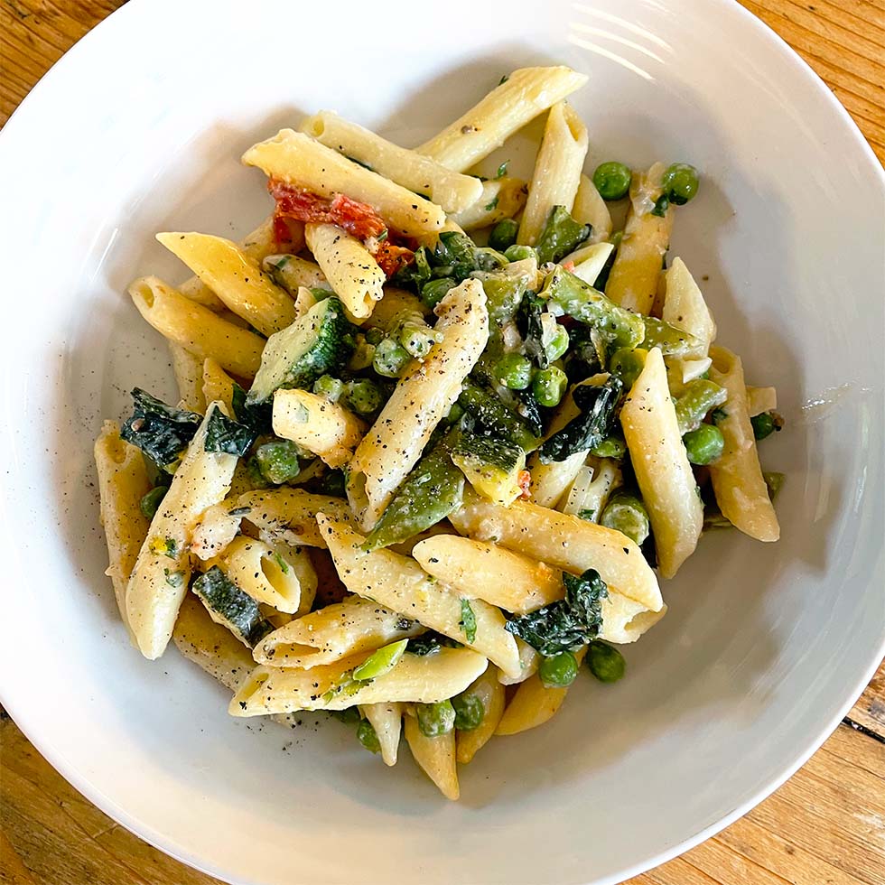 A white bowl filled with penne pasta mixed with green peas, leafy greens, and a creamy sauce, topped with cracked black pepper, sits on a wooden table.
