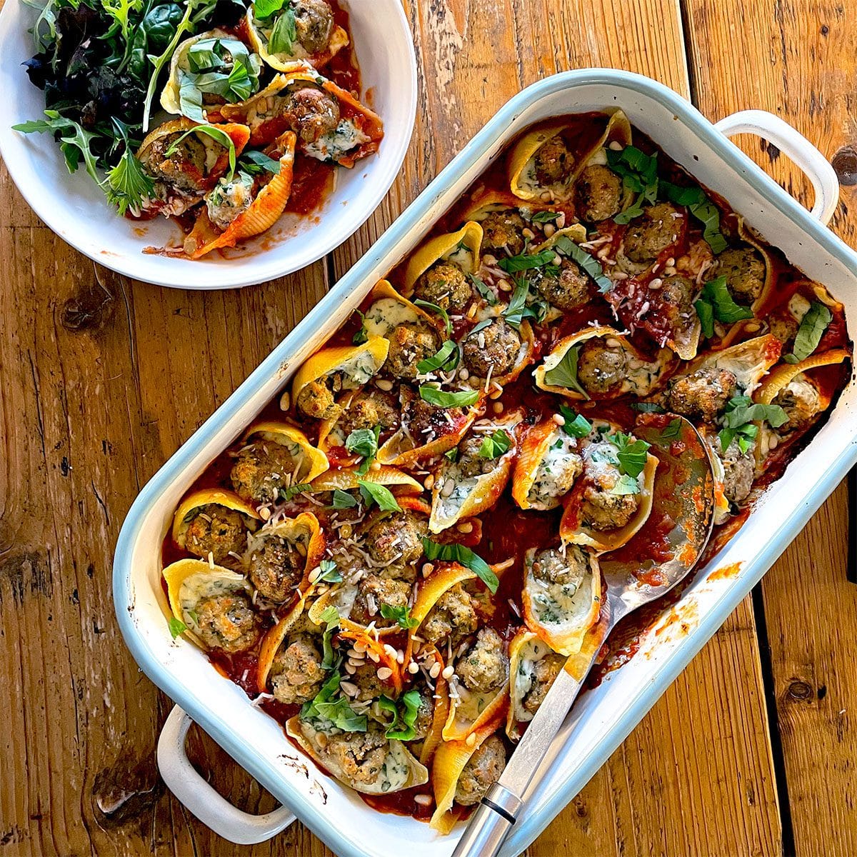 A baking dish filled with stuffed pasta shells topped with tomato sauce and herbs, with a serving spoon and a bowl of mixed green salad beside it on a wooden table.