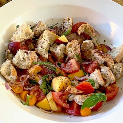 A bowl of panzanella salad with chunks of rustic bread, tomatoes, yellow cherry tomatoes, peach slices, red onion, fresh basil, and ground black pepper.