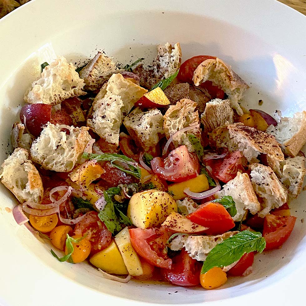 A bowl of panzanella salad with chunks of rustic bread, tomatoes, yellow cherry tomatoes, peach slices, red onion, fresh basil, and ground black pepper.