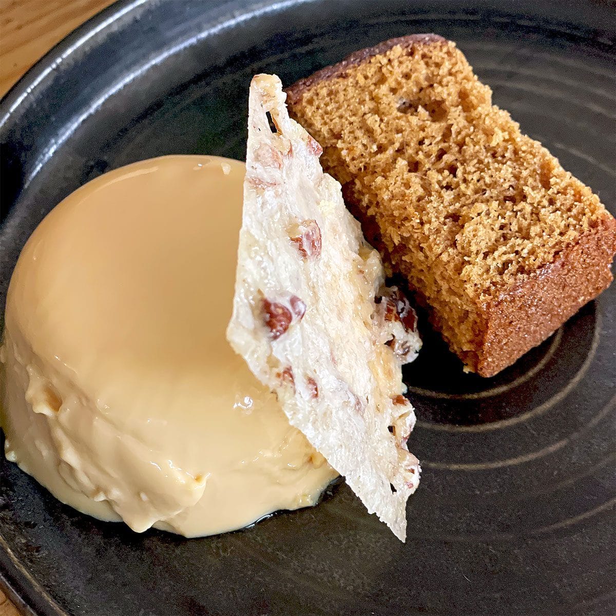 A round caramel-coloured dessert sits next to a slice of golden-brown cake, with a thin, textured tuile standing upright between them, all served on a dark ceramic plate.