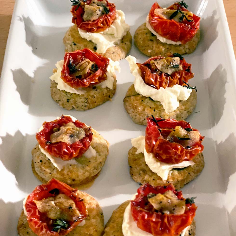 A white rectangular platter holds nine savoury appetisers made with a round biscuit base, topped with creamy cheese, roasted tomato halves, herbs, and small mushroom slices.