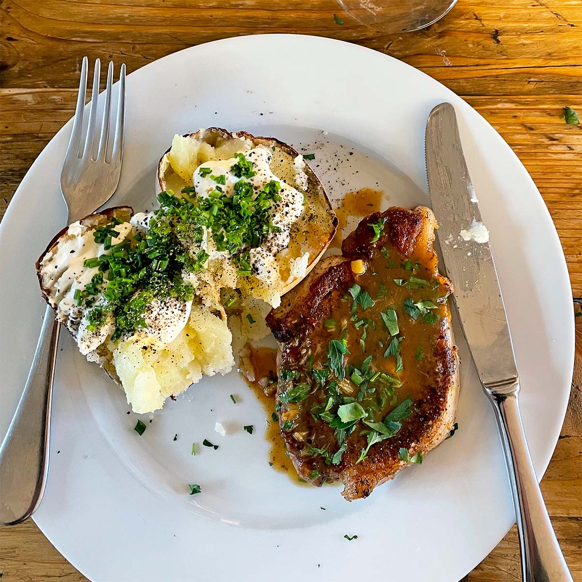 A white plate with a baked potato topped with soured cream, chopped chives, and pepper, next to a pork chop with brown sauce and herbs. A fork and knife are placed on the plate. The table is wooden.